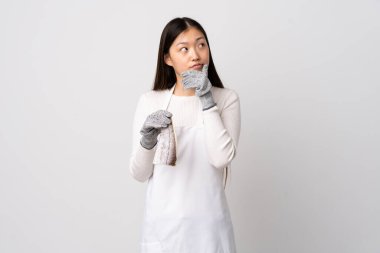 Çinli balıkçı önlük takıyor ve çiğ balığı izole edilmiş beyaz arka planda tutuyor. Şüpheleri var ve kafası karışmış bir yüz ifadesi var.