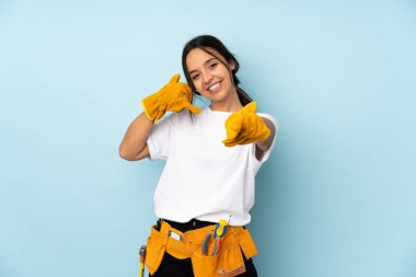 Genç elektrikçi kadın mavi arka planda izole edilmiş telefon hareketlerini yapıyor ve ön tarafı işaret ediyor.