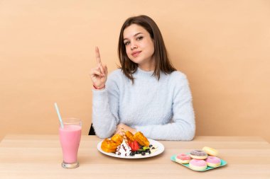 Bej arka planda izole edilmiş waffle yiyen genç kız en iyinin işareti olarak parmağını kaldırıyor.