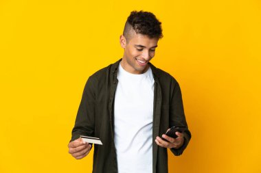 Genç Brezilyalı adam sarı arka planda izole edilmiş cep telefonuyla kredi kartıyla alışveriş yapıyor.