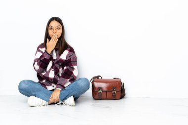 Genç bir kız öğrenci yerde oturuyor ve elleriyle ağzını kapatıyor.