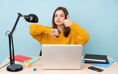 Öğrenci kız, mavi arka planda izole bir şekilde evinde ders çalışıyor. İyiye-kötüye işaret yapıyor. Evet ya da hayır arasında kararsız