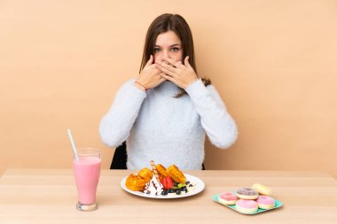 Bej arka planda izole edilmiş waffle yiyen genç kız ağızlarını elleriyle kapatıyor.
