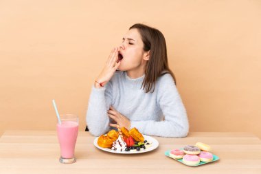 Bej arka planda izole edilmiş waffle yiyen genç kız esniyor ve ağzını eliyle açık tutuyor.