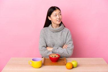 Genç Çinli bir kız bir masada kahvaltı edip gülümserken yukarı bakıyor.
