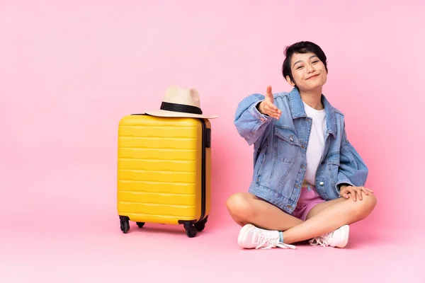 Young traveler Vietnamese woman with suitcase sitting on the floor over isolated pink background shaking hands for closing a good deal