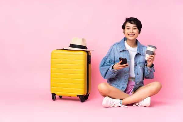 若いです旅行ベトナム人女性とともにスーツケース上の床の上に座って隔離されたピンクの背景保持コーヒーを取るためにと携帯電話 — ストック写真