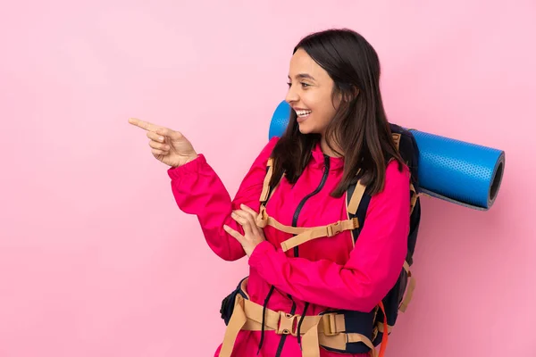 若いです登山家女の子とともに大きなバックパック上の隔離されたピンクの背景指差し指へと製品を提示 — ストック写真