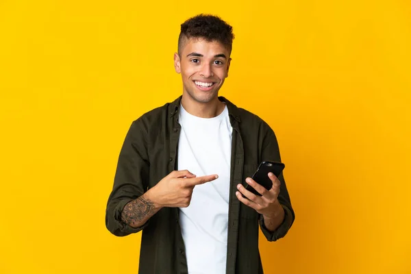 Junger Brasilianer Isoliert Auf Gelbem Hintergrund Mit Handy Und Zeigt — Stockfoto
