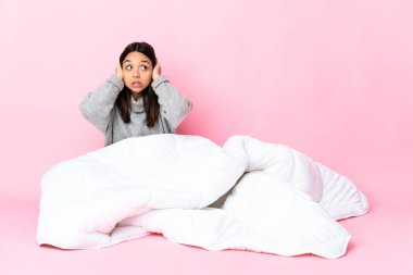 Genç melez kadın pijamasını giymiş yerde oturmuş sinirli ve kulaklarını tıkıyor.