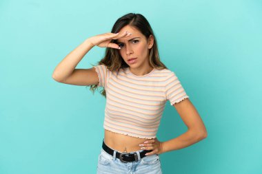 İzole edilmiş mavi arka planı olan genç bir kadın bir şeye bakmak için elini uzaklara bakıyor.