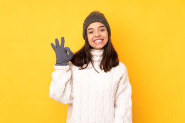 Sarı arka planda, kış şapkalı genç bir kadın parmaklarıyla tamam işareti gösteriyor.