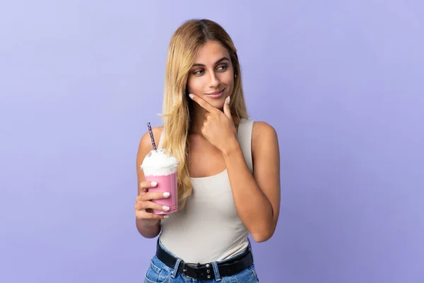 年轻的金发碧眼的乌拉圭籍女子 带着草莓奶昔 与紫色背景隔离 向侧面看去 面带微笑 — 图库照片
