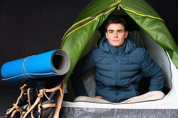 Teenager caucasian man inside a camping green tent isolated on black background angry