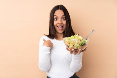 Genç esmer kız izole edilmiş bir arka planda salata tutuyor ve yüzünde sürpriz bir ifade var.