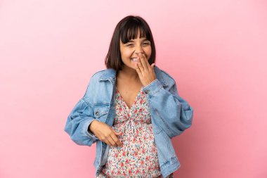 Genç hamile kadın pembe arka planda mutlu ve gülümsüyor elleriyle ağzını kapatıyor.