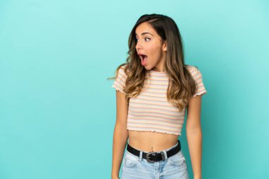 Soyutlanmış mavi arka planı olan genç bir kadın yan tarafa bakarken sürpriz jest yapıyor.