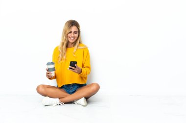 Genç sarışın Uruguaylı kadın, beyaz arka planda izole bir şekilde yerde oturuyor. Elinde kahve ve bir cep telefonu var.