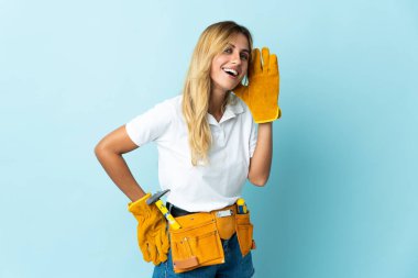 Genç sarışın Uruguaylı elektrikçi kadın mavi arka planda izole edilmiş bir şekilde elini kulağına koyarak bir şeyler dinliyor.