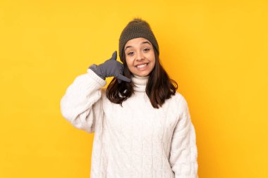 Sarı arka plan üzerinde kışlık şapkası olan genç bir kadın telefon hareketinde bulunuyor. Bana geri dönüş işareti ver.