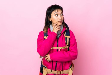 Pembe arka planda büyük bir sırt çantasıyla genç dağcı kız bir fikir düşünüyor.