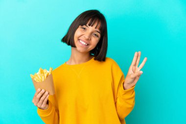 İzole edilmiş mavi arka planda patates kızartması tutan genç esmer kadın mutlu ve parmaklarıyla üçe kadar sayıyor.
