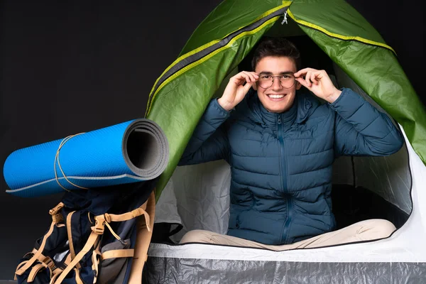 Teenager caucasian man inside a camping green tent isolated on black background with glasses and surprised