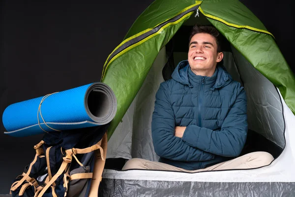 Teenager caucasian man inside a camping green tent isolated on black background looking up while smiling