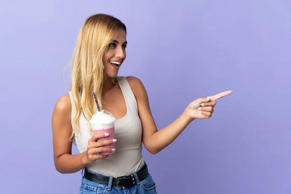 年轻的金发碧眼的乌拉圭女人 带着草莓奶昔 背景是紫色的 手指头指向旁边 并展示了一种产品 — 图库照片