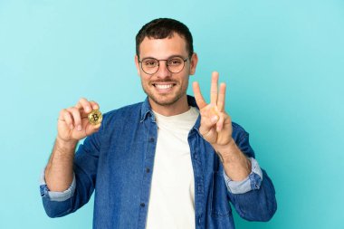 Brazilian man holding a Bitcoin over isolated blue background happy and counting three with fingers