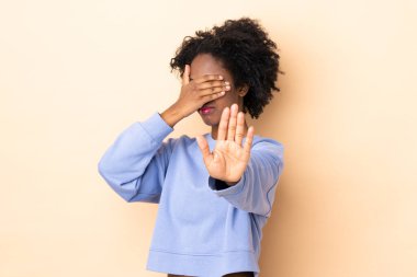 Genç Afro-Amerikalı kadın bej arka planda izole edilmiş. Durup jest yapıyor ve yüzünü kapatıyor.