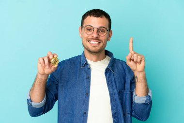 İzole edilmiş mavi arka plan üzerinde Bitcoin tutan Brezilyalı adam harika bir fikre işaret ediyor.