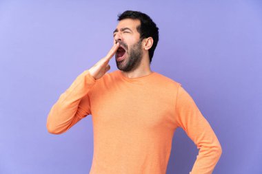 İzole edilmiş mor arka planda beyaz, yakışıklı bir adam esniyor ve ağzını eliyle kapatıyor.