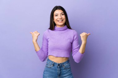 Genç esmer kadın, izole edilmiş mor arka planda, baş parmakları havada ve gülümsüyor.