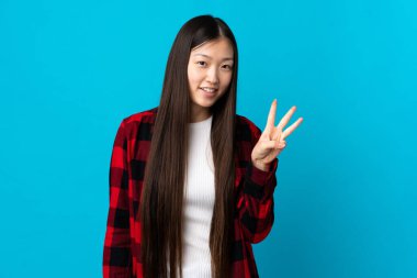 Soyutlanmış mavi arka planı olan genç Çinli kız mutlu ve parmaklarıyla üçe kadar sayıyor.