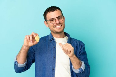 İzole edilmiş mavi arka planda Bitcoin tutan Brezilyalı adam gururlu ve kendini beğenmiş.