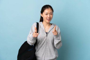 İzole edilmiş mavi arka planda spor çantalı genç Çinli kadın ön tarafı işaret ediyor ve gülümsüyor.