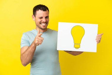 İzole edilmiş mor arka plan üzerinde Brezilyalı bir adam elinde ampul simgesi olan bir levha tutuyor ve ön tarafı işaret ediyor.