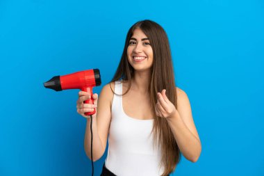Mavi arka planda saç kurutma makinesi tutan beyaz bir kadın para hareketi yapıyor.