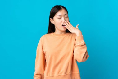 Genç Çinli kız, izole edilmiş mavi arka planda esniyor ve elleriyle ağzını kapatıyor.