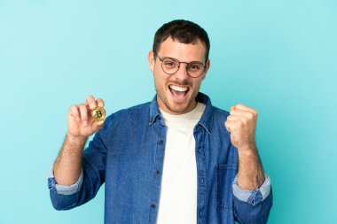 Elinde Bitcoin olan Brezilyalı bir adam mavi arka planda zaferi kutluyor.