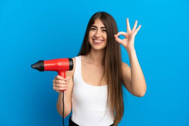 Mavi arka planda elinde saç kurutma makinesi tutan beyaz bir kadın parmaklarıyla tamam işareti gösteriyor.