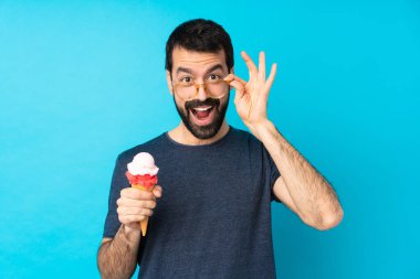 Kornet dondurması olan genç bir adam izole edilmiş mavi arka planda gözlüklü ve şaşırmış.
