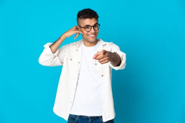 Genç Brezilyalı bir adam mavi arka planda izole edilmiş telefon hareketleri yapıyor ve ön tarafı işaret ediyor.