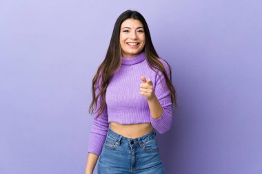 Genç esmer kadın, izole edilmiş mor arka planda şaşırmış ve ön tarafı işaret ediyor.