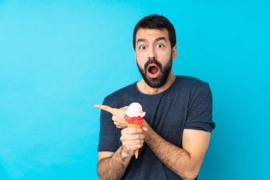 Kornet dondurması ile izole edilmiş mavi arka planı olan genç adam şaşırmış ve işaret ediyor.