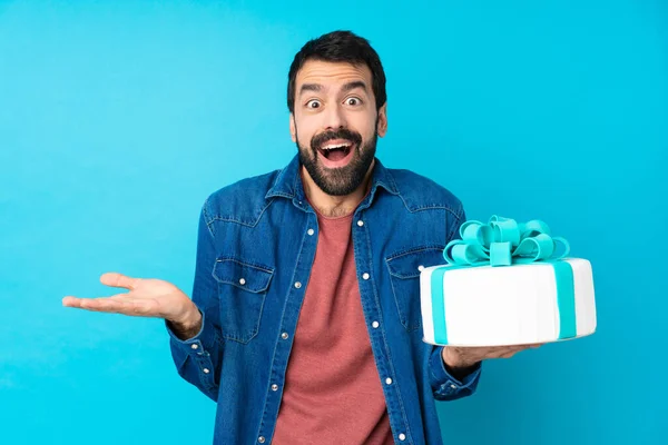 Young handsome man with a big cake over isolated blue background with shocked facial expression