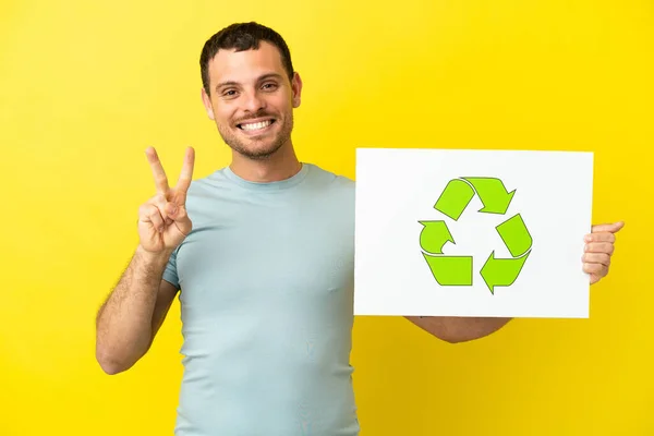 Brazylijczyk Nad Odizolowanym Fioletowym Tle Trzymając Tabliczkę Ikoną Recyklingu Świętując — Zdjęcie stockowe