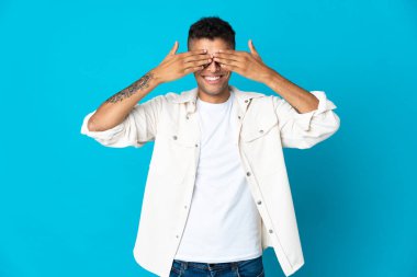 Genç Brezilyalı bir adam mavi arka planda izole edilmiş. Gözlerini elleriyle kapatıyor ve gülümsüyor.