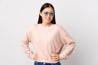 Genç Çinli kız izole edilmiş beyaz arka planda kolları kalçasında ve gülümsüyor.